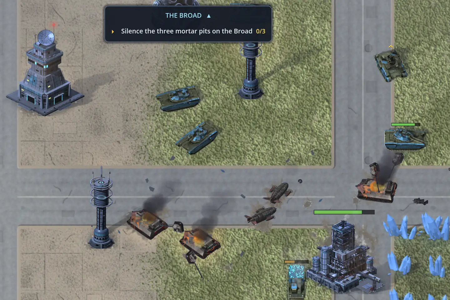 A campaign mission in a city, seen from above. A card at the top reads THE BROAD and “Silence the three mortar pits on the Broad 0/3”. Below it a crossroads is strewn with wreckage: two tanks burn and smoke on the road beside scattered scrap, olive tanks with blue trim move across the grass, a tall tower with a radar dish stands at the upper left beside two slim towers, and pale blue crystals glitter at the lower right.